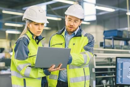 Male Industrial Worker and Female Chief Mechanical Engineer in Walk Through Manufacturing Plant while Discuss Factory's New Project and Using Laptop. Facility Has Working Machinery.