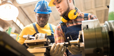 Low angle view of concentrated lathe operator wearing overall and hardhat distracted from work in order to answer questions of inspector carrying out quality control.