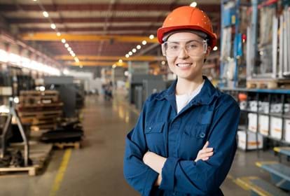 Young smiling female technician in blue uniform, protective eyeglasses and helmet working in modern factory