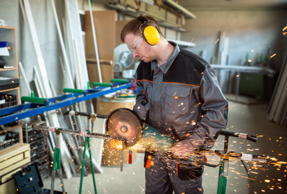 Worker cutting metal with grinder,while wearing protective gear.