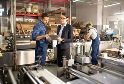 Two young confident workers in a processing factory discussing data on a tablet while standing by one of industrial machines
