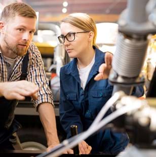 One of young confident technicians pointing at mechanism of industrial equipment during discussion of its qualities with colleague