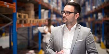 Successful businessman manager CEO holding tablet and walking through warehouse storage area looking towards shelves.