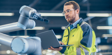 At the Factory: Automation Engineer Uses Laptop for Programming Robotic Arm. New Era in Automatic Manufacturing Industry.