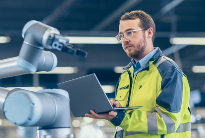 At the Factory: Automation Engineer Uses Laptop for Programming Robotic Arm. New Era in Automatic Manufacturing Industry.