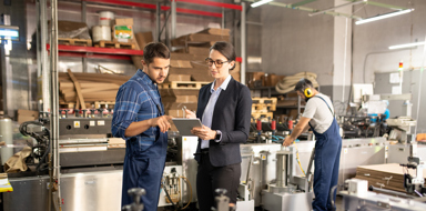 Two young confident workers in a processing factory discussing data on a tablet while standing by one of industrial machines